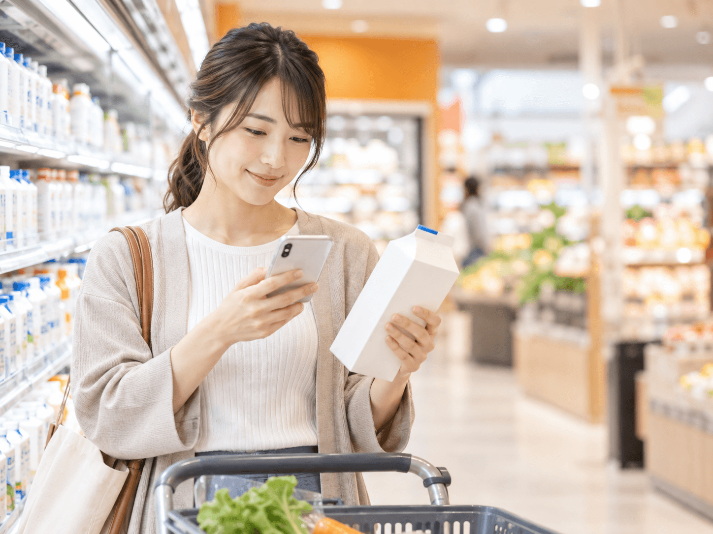 スーパーで商品を手に、スマートフォンで価格を確認する人